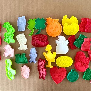 May include: A collection of colorful plastic cookie cutters in various shapes, including a pumpkin, ghost, shamrock, and Christmas tree. The cutters are in shades of red, green, yellow, pink, and blue.