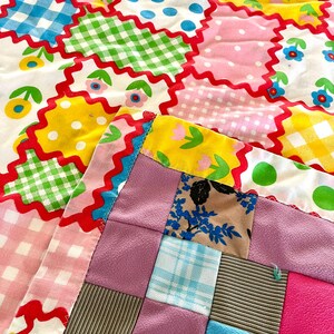 May include: A colorful patchwork quilt with a variety of patterns and colors. The quilt features squares of pink, blue, green, and yellow fabric with polka dots, gingham, and floral designs. The edges are trimmed with a red zigzag pattern.