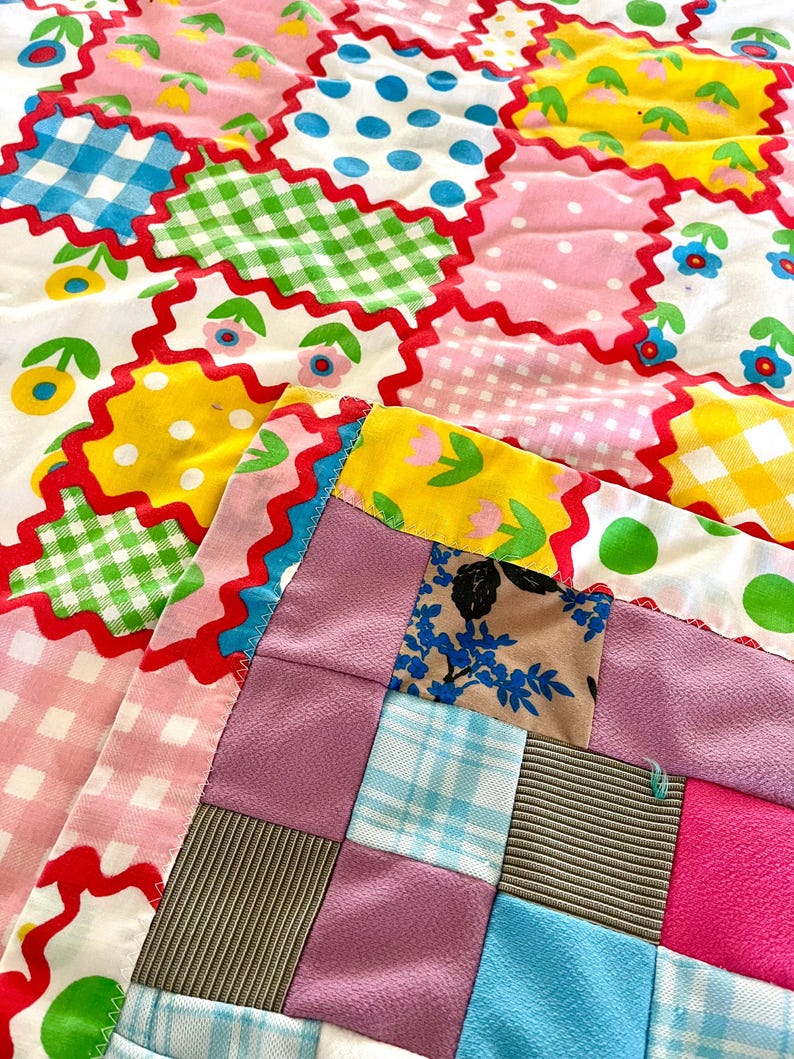 May include: A colorful patchwork quilt with a variety of patterns and colors. The quilt features squares of pink, blue, green, and yellow fabric with polka dots, gingham, and floral designs. The edges are trimmed with a red zigzag pattern.