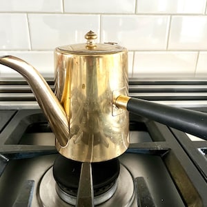 May include: A vintage brass coffee pot with a black handle and a long, curved spout. The pot is sitting on a gas stove burner. The pot has a lid with a small decorative knob.