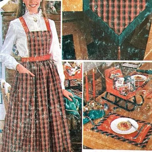 Op de afbeelding: Een vintage naaipatroon met een geruite schort en een bijpassende jurk. De schort heeft een rode tailleband en bandjes, met een volle rok. De afbeelding toont ook een tafelsetting met een bord eten en een kleine rode slee.