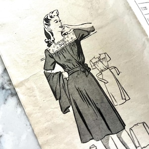 Könnte beinhalten: Vintage Schnittmuster mit detaillierter Illustration einer Frau in einem Kleid mit Gürtel und weiten Ärmeln. Das Muster enthält eine kleinere Skizze des Kleides und Text mit Größenangaben. Das Bild ist in Schwarzweiß.