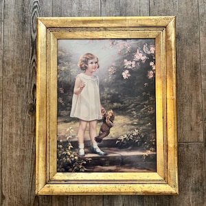 Puede incluir: Una impresión vintage enmarcada que presenta a un niño pequeño con un vestido blanco, sosteniendo un animal de peluche. La obra de arte está ambientada en un jardín con elementos florales y un marco dorado ornamentado.