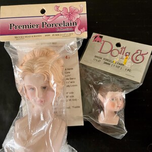 May include: Two porcelain doll heads in plastic packaging. The larger head is 8.9 cm and has blonde hair. The smaller head is 3.8 cm and has brown hair. The packaging includes the text "Premier Porcelain Collection" and "Brooke Head & Hands".