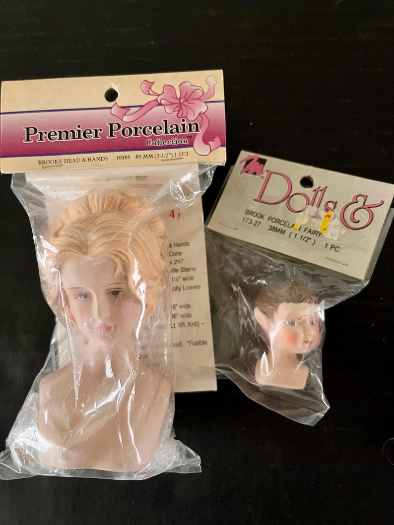 May include: Two porcelain doll heads in plastic packaging. The larger head is 8.9 cm and has blonde hair. The smaller head is 3.8 cm and has brown hair. The packaging includes the text "Premier Porcelain Collection" and "Brooke Head & Hands".