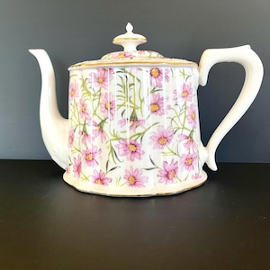 Op de afbeelding: Witte keramische theepot met een deksel, versierd met roze madeliefbloemen en groene bladeren. De theepot heeft een gebogen tuit en handvat, en een gouden rand. Een decoratief stuk voor het serveren van thee, met een inhoud van ongeveer 1,2 liter.