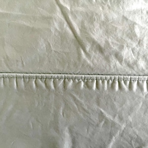 Könnte beinhalten: Nahaufnahme eines Stoffabschnitts, der horizontal durch eine Naht geteilt ist. Der Stoff hat eine leicht zerknitterte Textur und eine gedämpfte, silbrig-grüne Farbe. Die Naht ist mit dunklerem Faden genäht.