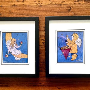 May include: Two framed art prints featuring whimsical angel illustrations. One shows an angel on a crescent moon holding a rose, the other depicts an angel with an umbrella catching stars. Both have a blue background with white stars.