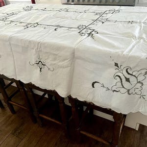 May include: A rectangular white tablecloth featuring black embroidered floral motifs and a central rectangular border design. The fabric has a scalloped edge finish along the perimeter, showcasing detailed needlework patterns across the surface.