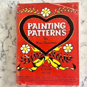 以下が含まれることがあります： ルース・ワイエス・スピアーズ著の「PAINTING PATTERNS for Home Decorators」というタイトルのヴィンテージ本。表紙は赤色で、タイトルが白で書かれた黒いハート型のデザインです。表紙には花柄とリボンのイラストが飾られており、内容を詳しく説明するテキストが添えられています。
