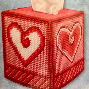 Può includere: Un copri scatola di fazzoletti fatto a mano, nei toni del rosso e del rosa, con un grande motivo a cuore su due lati. La scatola è quadrata e ha un'apertura superiore per i fazzoletti.