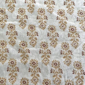 May include: A white fabric featuring a repeating block-printed pattern of stylised floral motifs in shades of golden yellow and brown. The design consists of small flowers with central brown dots and leafy stems arranged in a consistent grid-like layout.