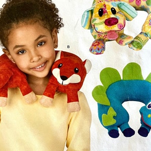 May include: A child wears a red fox-shaped neck pillow. Other neck pillows in the image include a dog with floral patterns and a blue dinosaur. These plush pillows are designed for comfort and support.