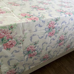 May include: A floral patterned bedspread featuring large pink roses, green leaves, and light blue ribbons on a cream background. The bedspread is draped over a bed frame, showcasing the full pattern. The overall theme is vintage and romantic.