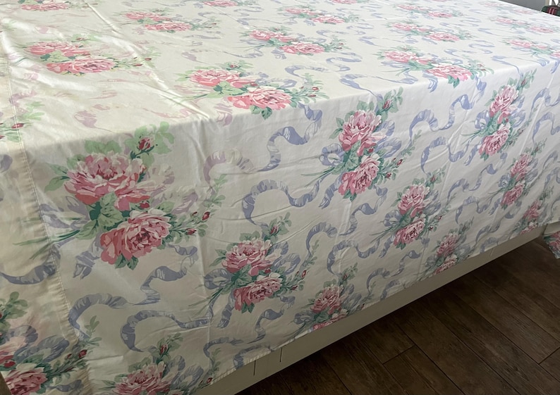 May include: A floral patterned bedspread featuring large pink roses, green leaves, and light blue ribbons on a cream background. The bedspread is draped over a bed frame, showcasing the full pattern. The overall theme is vintage and romantic.