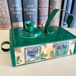 May include: A rectangular green metal candle holder featuring a curved handle and a small circular side loop. The front panel displays two framed illustrations of a moose and a cabin in a forest setting, surrounded by a decorative leaf pattern.