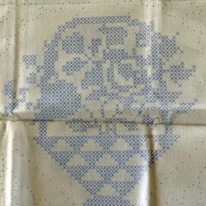 Könnte beinhalten: Cremefarbener Stoff mit einem großen, zentralen Kreuzstichmuster in Blau. Das Design scheint ein stilisiertes Blumen- oder geometrisches Muster zu sein. Der Stoff hat eine subtile gepunktete Textur.