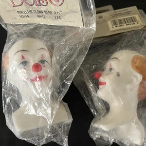 May include: Two porcelain clown heads, one with a red nose and a white face, and the other with a brown wig and a red nose. Both heads are 3 3/4 inches tall and are packaged in plastic bags.