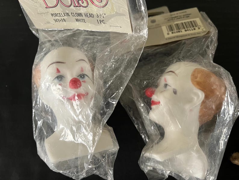 May include: Two porcelain clown heads, one with a red nose and a white face, and the other with a brown wig and a red nose. Both heads are 3 3/4 inches tall and are packaged in plastic bags.