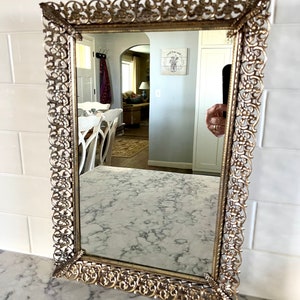 Puede incluir: Un espejo adornado de color dorado con un marco de filigrana decorativo. El espejo refleja una habitación con paredes blancas y una mesa blanca con sillas.
