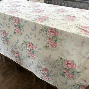 May include: A rectangular tablecloth with a floral pattern. The tablecloth features a cream background with pink peony bouquets and light purple ribbon accents. The tablecloth is draped over a wooden table.