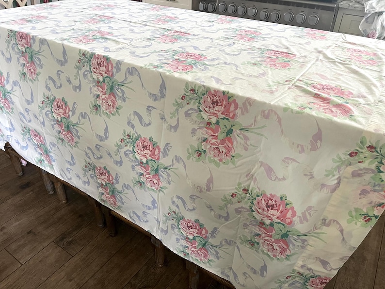 May include: A rectangular tablecloth with a floral pattern. The tablecloth features a cream background with pink peony bouquets and light purple ribbon accents. The tablecloth is draped over a wooden table.