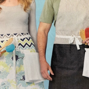May include: Two aprons, one with a chevron pattern and the other with a solid black bottom and a striped top. Both aprons have a white towel attached to the side and a wooden spoon and spatula.