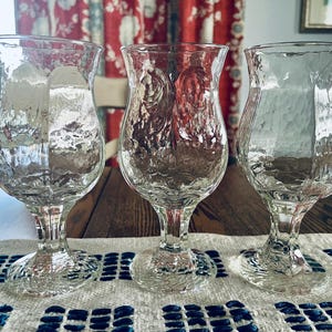 May include: Three clear, textured glass goblets with a tulip-shaped bowl and a stemmed base. The goblets are arranged on a white and blue patterned runner on a wooden table. The background includes a window, a chair, and red floral curtains.