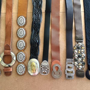 May include: A selection of belts in different styles and colours. The belts include black, brown, and tan leather, some with decorative buckles. The buckles feature silver and gold tones, with some having intricate designs and embellishments.
