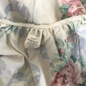 May include: A close-up of a Queen-sized, floral-patterned bed sheet. The sheet features a cream-colored background with a design of pink roses, green leaves, and blue ribbons. The tag reads "Queen No Iron Percale".