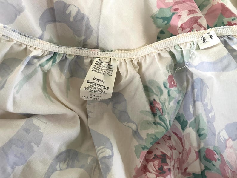May include: A close-up of a Queen-sized, floral-patterned bed sheet. The sheet features a cream-colored background with a design of pink roses, green leaves, and blue ribbons. The tag reads "Queen No Iron Percale".