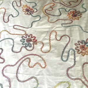 Può includere: Tessuto bianco con ricami floreali e a catena colorati. Il ricamo presenta una varietà di colori, tra cui rosso, arancione, giallo, blu e rosa. I disegni floreali sono raggruppati e i disegni a catena si snodano sul tessuto.