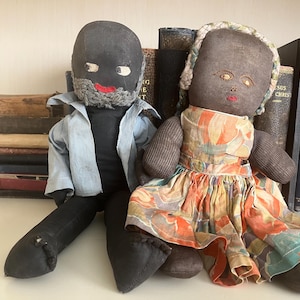 May include: Two handmade cloth dolls. One doll has a black head, gray beard, and wears a blue jacket. The other doll has a brown head and wears a colorful dress. They are sitting in front of a collection of antique books.
