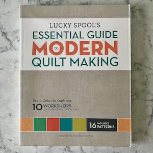 May include: A book titled "Lucky Spool's Essential Guide to Modern Quilt Making" with text in teal and orange. The cover includes the text "From Color to Quilting: 10 Workshops by Your Favorite Teachers" and "16 Patterns". Compiled by Susanne Woods.