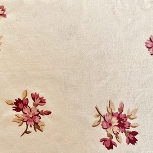 Puede incluir: Una tela de color crema con un patrón repetitivo de pequeños grupos florales. Cada grupo consta de flores rosas y magenta intenso con hojas y tallos de color tostado claro, distribuidos uniformemente sobre la superficie textil lisa.