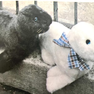Può includere: Due foche di peluche, una nera e una bianca. La foca nera ha occhi blu e baffi. La foca bianca presenta occhi blu, un naso nero, baffi e un nastro a quadri blu, marrone e bianco legato attorno al collo con un fiocco.