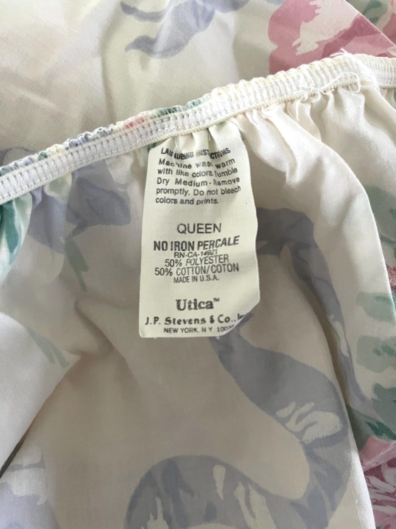 May include: Close-up of a floral patterned Queen size bed sheet. The sheet features a white background with pink, blue, and green floral designs. A tag with washing instructions and the brand "Utica" is visible.