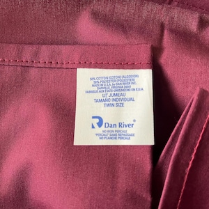 May include: Close-up of a burgundy-colored fabric with a sewn seam and a white label. The label reads "Twin Size" and includes the Dan River logo. The fabric is a blend of cotton and polyester.