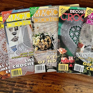 May include: A collection of vintage "Magic Crochet" magazines, featuring various crochet patterns. The covers display intricate crochet designs, floral arrangements, and text such as "Gift Giving Crochet" and "Christmas Decor". The magazines are arranged on a wooden surface.