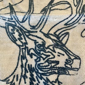 Può includere: Primo piano di un tessuto con uno schizzo blu scuro della testa e delle corna di un cervo. Il disegno è su un tessuto color crema, strutturato, che mostra il lavoro di linea dettagliato dell'illustrazione.