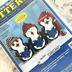 Könnte beinhalten: Eine Packung mit einem Holzbearbeitungsmuster für ein Trio singender Pinguine. Die Pinguine sind mit Hüten und Schals dargestellt und halten Gesangbücher. Die Packung enthält Anweisungen für eine 55.88 cm hohe Gartenausstellung.