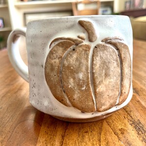 Könnte beinhalten: Eine handgefertigte Keramiktasse mit einem Kürbismotiv. Die Tasse ist weiß mit einem braunen Kürbisrelief. Die Tasse hat einen Henkel und steht auf einer Holzoberfläche. Die Tasse ist etwa 10 cm hoch.