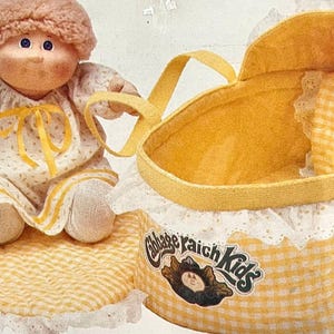 Può includere: Una bambola con capelli in lana marrone chiaro che indossa un vestito bianco con motivo floreale giallo e nastro giallo, accanto a una culla in tessuto a quadretti gialli e bianchi con bordo in pizzo bianco e logo Cabbage Patch Kids.