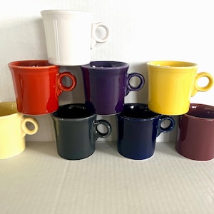 Puede incluir: Un conjunto de nueve tazas de cerámica en varios colores: blanco, rojo, morado, amarillo, negro, azul marino y granate. Cada taza tiene un asa redondeada y un borde ligeramente acampanado.