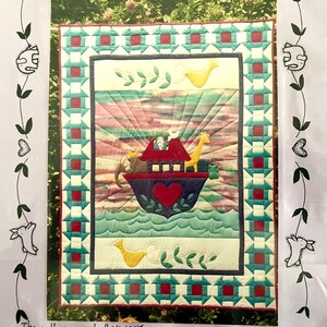 Pode incluir: Uma colcha colorida com uma borda de patchwork e um painel central que representa uma arca vermelha e azul com animais em um mar azul e verde. A arca tem um coração vermelho no lado. O fundo é um pôr do sol rosa e roxo.