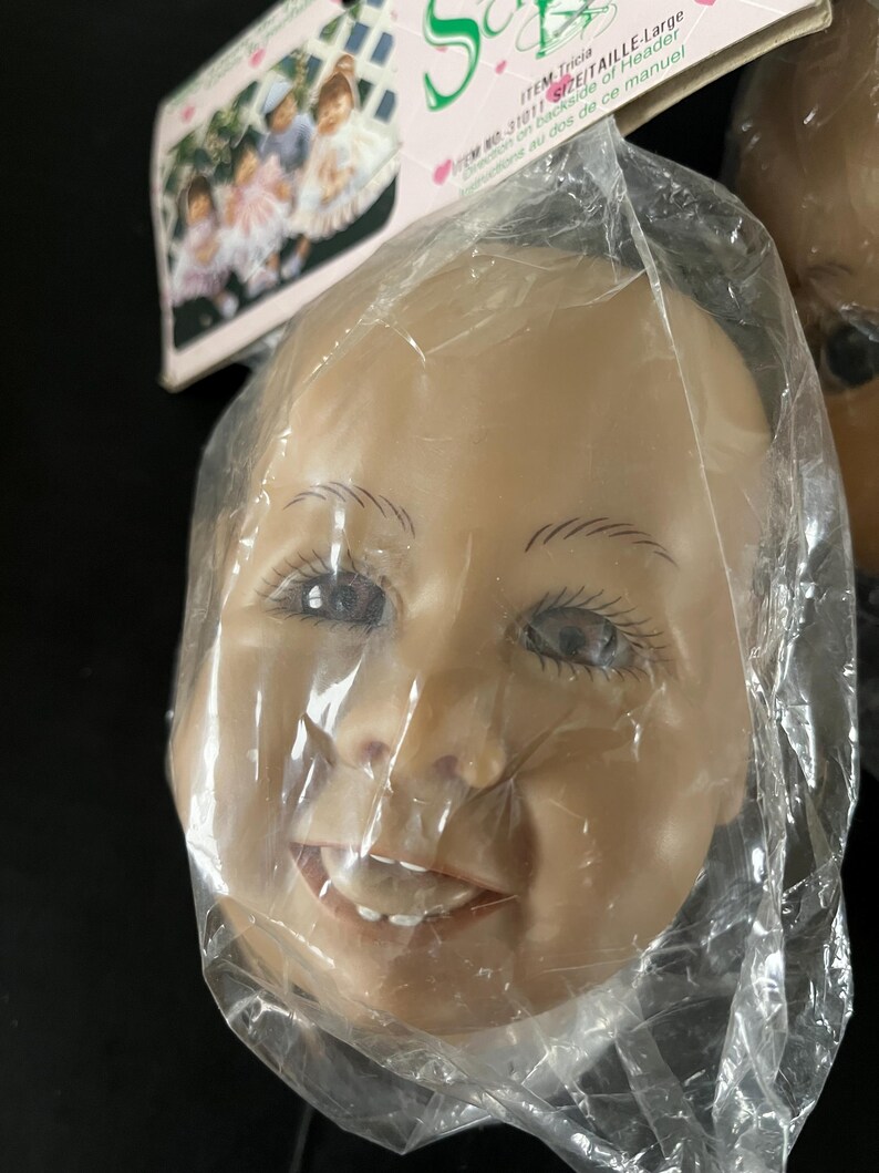 May include: Close-up of a doll's head with a smiling expression. The head is made of a realistic-looking material and has painted features, including brown eyes, eyebrows, and a mouth with teeth. The head is packaged in a clear plastic bag.