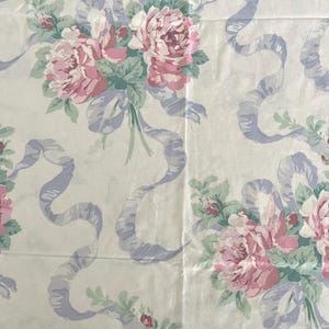 May include: Cream-colored fabric with a repeating pattern of pink roses, green leaves, and light purple ribbons. The floral design is arranged in bouquets, creating a vintage or romantic aesthetic. The fabric appears to be cotton or a similar material.
