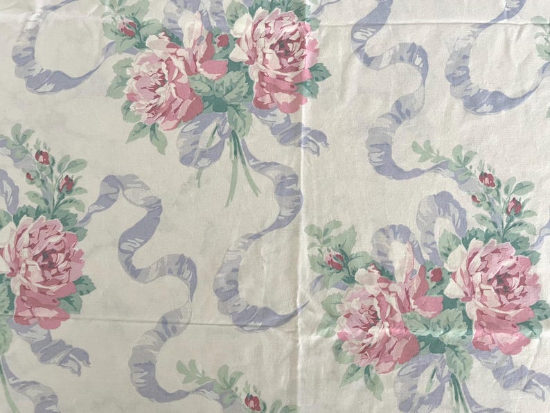 May include: Cream-colored fabric with a repeating pattern of pink roses, green leaves, and light purple ribbons. The floral design is arranged in bouquets, creating a vintage or romantic aesthetic. The fabric appears to be cotton or a similar material.