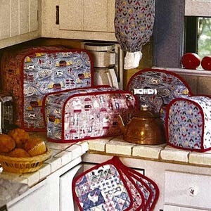 May include: A collection of kitchen accessories, including toaster covers, a kettle cover, and pot holders. The covers feature a patterned fabric with red trim. A copper kettle and a basket of muffins are also visible.
