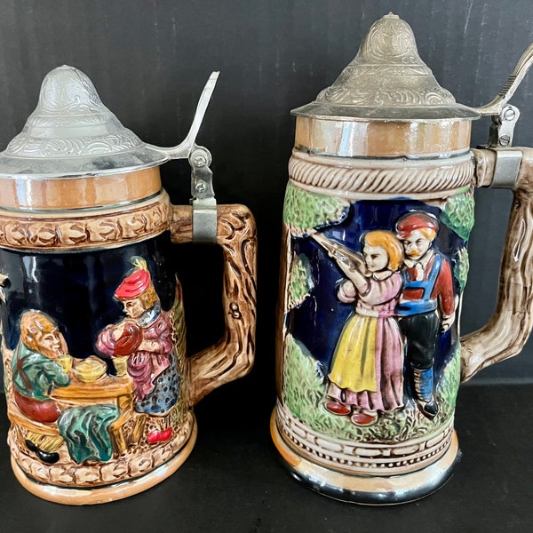 Vintage German beer steins with attached metal lids and hand-painted, raised relief exteriors, made in Japan. One 7" tall, one 7.75" tall.